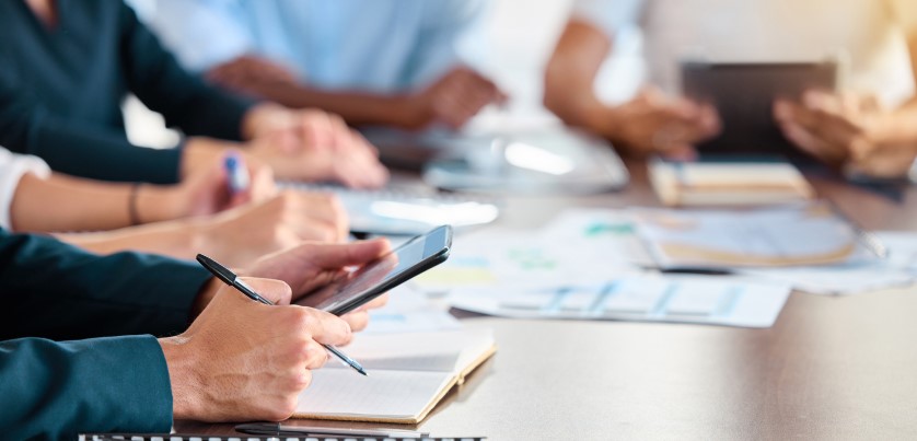 people sat around a desk with tablet computers, phones, paperwork and writing pads